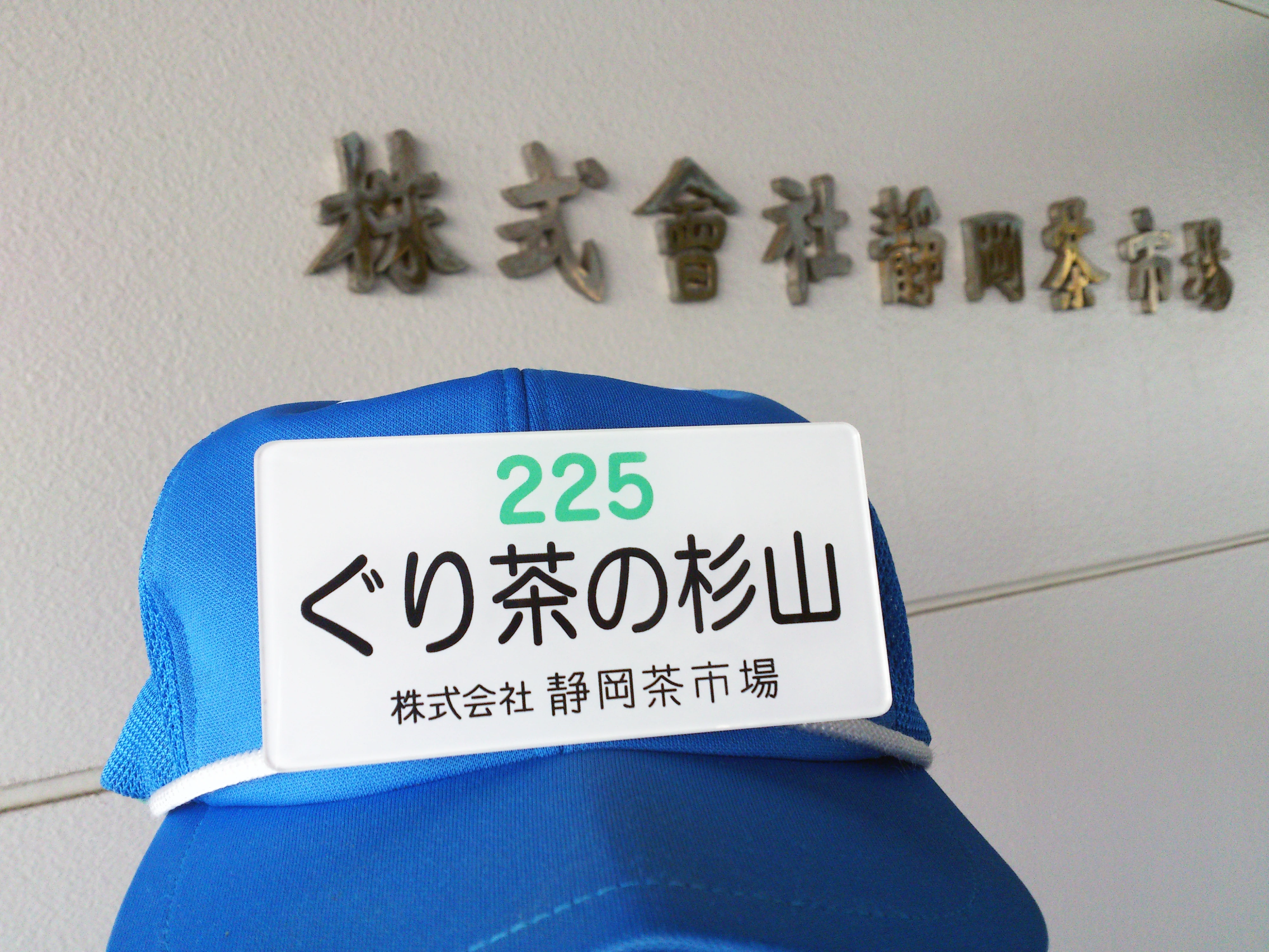 静岡茶市場の建物を背景にした、ぐり茶の杉山の名札と帽子